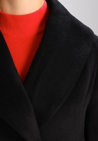 Schwarzer, strukturierter Mantel mit breitem Revers, über einem roten, gestreiften Rollkragenpullover getragen. Der Mantel hat eine weiche, flauschige Oberfläche.