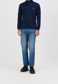 Marinefarbener Rollkragenpullover mit langen Ärmeln, aus weichem Stoff, kombiniert mit hellblauen geraden Jeans und schwarzen Sneakers. Kleines Logodetail auf der Brust.