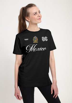 Kvinde med hestehale iført sort T-shirt med teksten "Mexico", nummer 86 og farverigt emblem, stående mod ensartet lyst baggrund.
