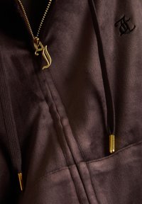Donkerbruin suède jack met een gouden ritssluiting in de vorm van een gestileerde letter "J" en bijpassende gouden trekkoorden.
