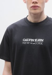 Melna kokvilnas T-krekls ar apaļu kakla izgriezumu, uz kura ir izšūts teksts "CALVIN KLEIN NEW EST. 1968 YORK" baltā krāsā. Gluda tekstūra un ērtas piegriezums.