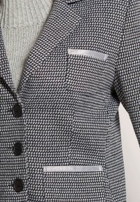 Blazer mit einem strukturierten schwarz-weißen Hahnentrittmuster, mit fünf Knöpfen und einer silberfarbenen Tasche auf der Brust.