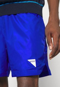 Pantaloni da bagno blu realizzati in un tessuto leggero e strutturato. Presentano una piccola toppa con logo bianco e un motivo a griglia; vita elasticizzata per una vestibilità ottimale.