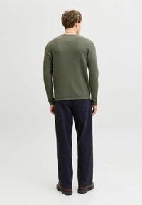 Man standing facing away wearing olive green sweater, dark navy wide-leg pants, and brown shoes on white background.