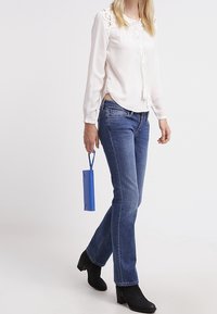 Blouse blanche à manches longues avec des accents en dentelle, associée à un jean bleu et des bottines noires, tenant un petit sac bleu.