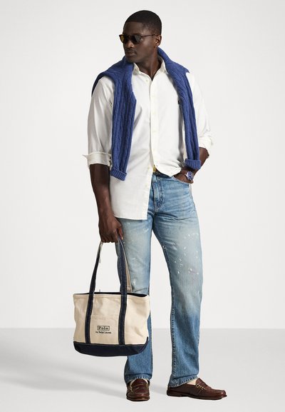 Beżowa i granatowa torba z płótna z podwójnymi uchwytami, o prostokątnym kształcie i z logo. Model nosi białą koszulę, niebieski sweter i dżinsy.