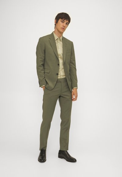 Olivgrüner Anzug mit taillierter Passform, einreihiger Blazer und passender Hose, kombiniert mit einem hellbeigen Hemd und schwarzen Schuhen.