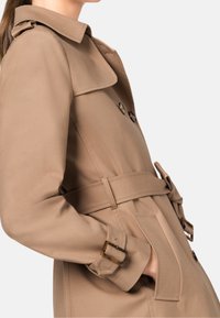 Trench-coat beige avec boutons croisés, taille ceinturée, grand col et poignets ajustables. Tissu lisse avec une coupe cintrée.