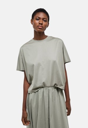 Femme debout portant une chemise ample à manches courtes gris clair et une jupe taille haute assortie, sur fond blanc.