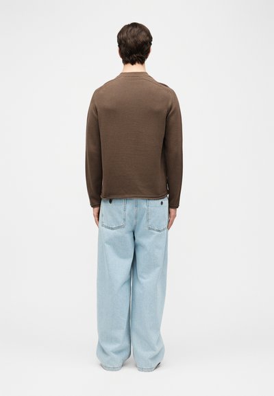 Άνδρας που φοράει καφέ πουλόβερ με μακριά μανίκια και φαρδιά ανοιχτό μπλε τζιν, όρθιος με την πλάτη γυρισμένη προς ένα απλό λευκό φόντο.