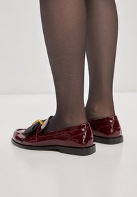 Mocassini in pelle verniciata bordeaux con un accentuato fiocco, hardware dorato e una suola nera bassa, indossati con collant neri trasparenti.