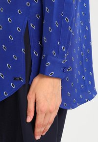 Camicia blu con una superficie liscia, caratterizzata da un motivo geometrico bianco. Colletto dritto e polsini abbottonati con una fessura laterale per un tocco in più.