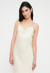 Φόρεμα slip σε κρεμ χρώμα με ντεκολτέ καστόρι, λεπτές τιράντες και ζώνη δετή στη μέση, κατασκευασμένο από ελαφρύ, λείο ύφασμα.
