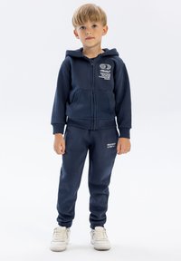 Junger Junge steht auf, trägt einen navyblauen Hoodie und passende Jogginghosen mit weißen Turnschuhen, vor einem einfarbigen hellen Hintergrund.
