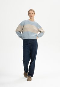 Junge Frau mit blonden Haaren, die einen blau-beigen gestreiften Pullover, dunkle Jeans und braune Schuhe trägt und mit den Händen in den Taschen steht.