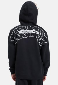 Čierna mikina s kapucňou obsahujúca veľké biele grafické logo na chrbte, s rebrovanými manžetami a lemom. Vyrobená z jemnej, textúrovanej látky.