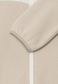 Gros plan sur une veste polaire beige avec une fermeture éclair visible à l'avant et un poignet élastique sur la manche, montrant un tissu doux et texturé.