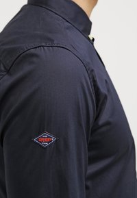 Námořnická modrá košile na knoflíky vyrobená z hladkého materiálu. Má stojací límec a diamond tvarovaný nášivku "Superdry" na levém rukávu.