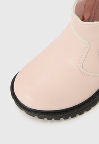 Gros plan sur une botte de cheville rose clair avec une surface brillante et une semelle en caoutchouc noir, mettant en avant les coutures et les détails de design.