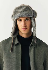 Hallus hallist faux karusnahast müts kõrva katetega, mille külgedel on ruuduline muster ja nupuvajutus. Kandmine musta kõrge kaelusega särk ja roheline jakk.