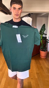 Camiseta de algodón verde con mangas cortas, que presenta un diseño de ballena bordado en el lado izquierdo del pecho, con etiqueta adjunta. Fondo de suelo de madera.