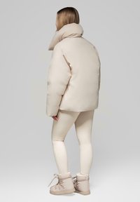 Beige péřová bunda s vysokým límcem, ve spojení se světlými béžovými legínami a odpovídajícími kotníkovými botami, které mají hladký, texturovaný povrch.