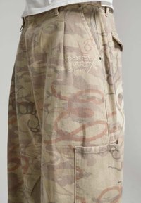 Pantalon cargo beige avec des motifs abstraits de graffiti rouge et marron délavés, avec des plis et une poche latérale sous une chemise blanche.