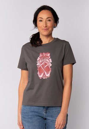 Vrouw met donker haar in een paardenstaart, gekleed in een grijs T-shirt met roze balletpantoffels en een bloemdessin, gecombineerd met een blauwe jeans.