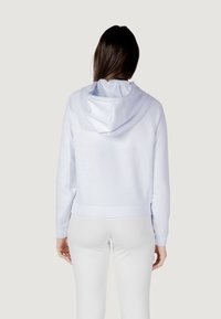 Cropped hoodie azzurro chiaro realizzato in tessuto morbido, con cappuccio regolabile e orlo a coste. Indossato con leggings bianchi, che mostrano una texture liscia.