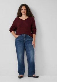 Bordeaux gebreide trui met een V-hals en opgerolde mouwen, gecombineerd met blauwe straight-leg spijkerjeans. Zwarte platte schoenen maken de outfit compleet.