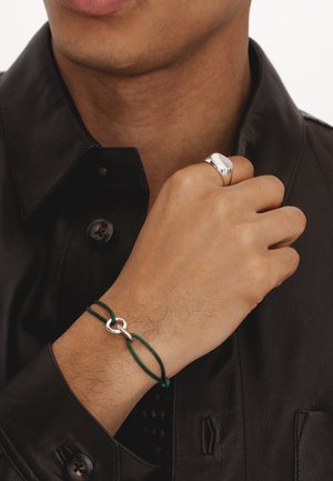 Zaļš corda rokassprādze ar sudraba ovālu saiti. Valkāta uz rokas, papildināta ar sudraba gredzenu un melnu ādas krekli.