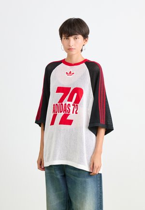 Junge Person mit kurzen Haaren, die ein weißes Adidas-Jersey aus Netzstoff mit roten und schwarzen Ärmeln sowie lockeren blauen Jeans trägt und vor einem weißen Hintergrund steht.