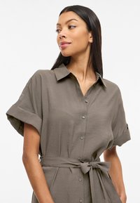 Γυναίκα που φορά γκρι κοντομάνικο φόρεμα με κουμπιά, γιακά και δεμένη ζώνη στη μέση, κοιτάζει προς τα αριστερά σε λευκό φόντο.