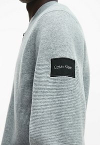 Zbliżenie na ramię osoby noszącej jasnoszary sweter dzianinowy z czarną naszywką logo Calvin Klein na górnej części ramienia.