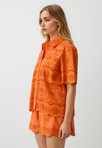 Oranje blouse met oogjes, een kraagnek, korte mouwen en bloemen uitsparingen; gecombineerd met bijpassende shorts met een scalloped zoom.