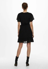 Vestito nero con maniche corte trasparenti a sbuffo, orlo svasato con rouches, dettaglio con cintura in vita, indossato con stivali neri alla caviglia.