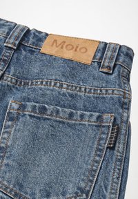 Džins hlače s modrim pranjem, ki imajo bež usnjeno oznako s pritisnjenim napisom "Molo." Vključujejo kontrastno oranžno šivanje in majhno črno oznako.