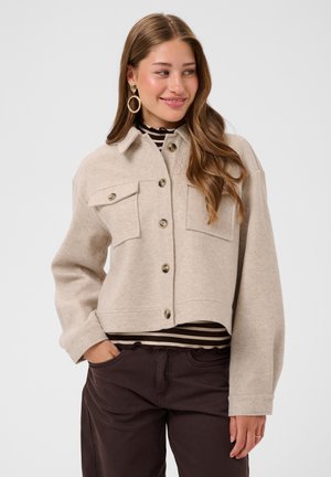 Ung kvinde med langt hår iført beige knapjakke over stribet top og mørke bukser, smilende og ser til siden.