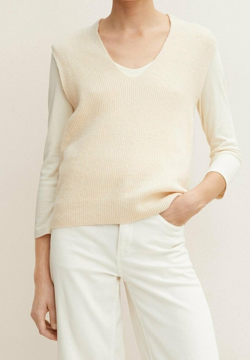 Beige ribbad stickad tröja med djup V-hals, lager på lager över en krämfärgad långärmad topp, bärs med vita byxor. Mjuk textur och avslappnad stil.