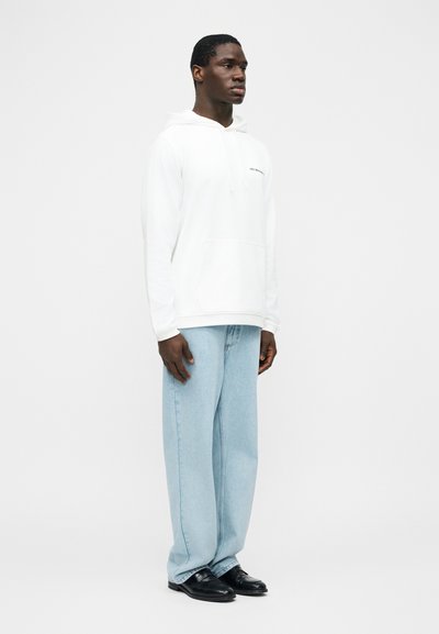 Hombre de pie con una sudadera blanca con texto, jeans anchos de mezclilla azul claro y zapatos de cuero negros frente a un fondo blanco liso.