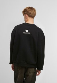 Svart oversized sweatshirt med rund hals og senket skuldre. Har hvit "UPSCALE STUDIO" tekst og logo på baksiden.