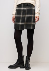 Zwarte geruite rok met beige accenten, voorzien van een tailleband met gesp en plooi-details, gecombineerd met zwarte panty's en enkellaarsjes.