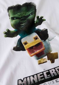 T-skjorte med en illustrasjon av en grønn figur med svart hår som rir på en hvit and, på en hvit stoffbakgrunn. Teksten under lyder «A Minecraft Movie.»