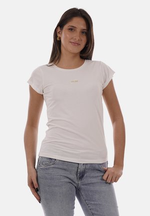 Jeune femme aux longs cheveux foncés, portant des boucles d'oreilles créoles dorées, un t-shirt blanc ajusté avec un petit logo doré et un jean gris clair, debout avec une main dans la poche.