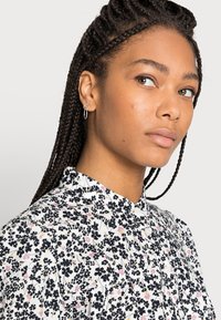 Blusa branca com padrão floral em preto e rosa, com gola e pala de botões. O modelo tem cabelo trançado e brincos de argola.