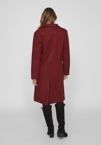 Manteau long bordeaux avec un col cranté, une fente arrière simple et un tissu texturé. Porté avec des bottes noires à talons hauts.