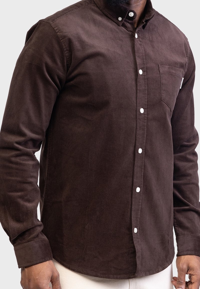 Brązowa koszula z korduroyu z kołnierzykiem zapinanym na guziki, długimi rękawami i kieszonką na piersi. Posiada białe guziki i delikatną strukturę.