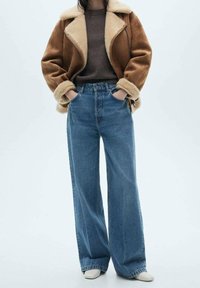 Veste en daim marron avec doublure en peau lainée beige, associée à un jean bleu taille haute à jambes larges et des chaussures de couleur claire.