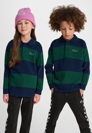 Gestreifte langärmelige Poloshirts in Grün und Marineblau mit Kragen, kombiniert mit schwarzen Hosen mit weißen Seitenstreifen; eine Person trägt eine rosa Strickmütze.