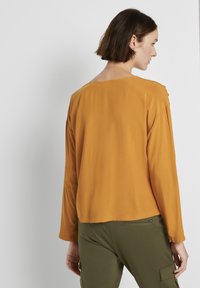 Donna con i capelli corti castani che indossa un top a maniche lunghe senape e pantaloni verde oliva, in piedi di lato contro uno sfondo bianco semplice.